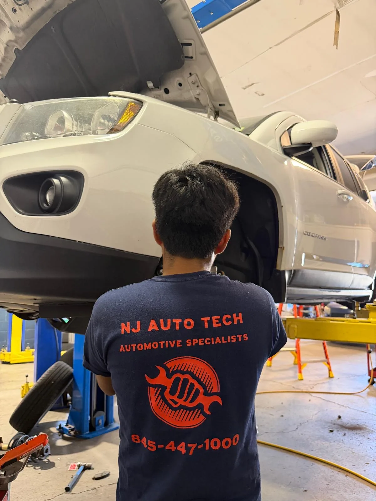 NJ Auto Tech technician inspecting the front end of a white SUV with the hood open and wheel removed